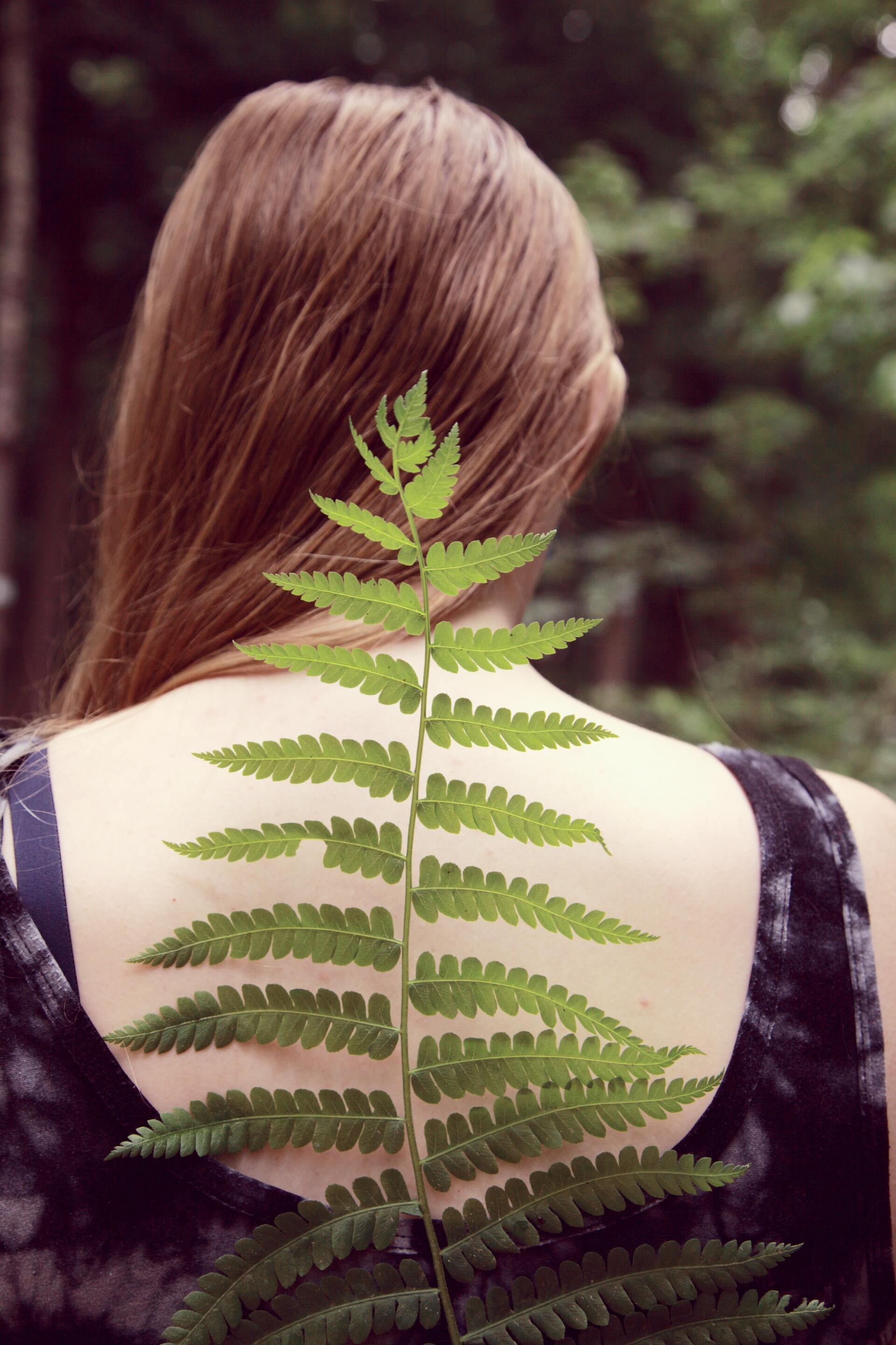 Woman with long brown hair, wearing a dark dress, holds a green fern frond on her back in a forest.